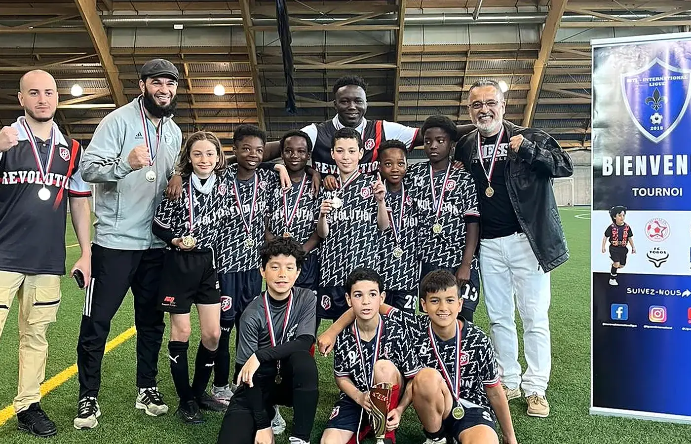 Équipe Révolution médaillée posant avec ses encadrants devant la bannière d'accueil d'un tournoi